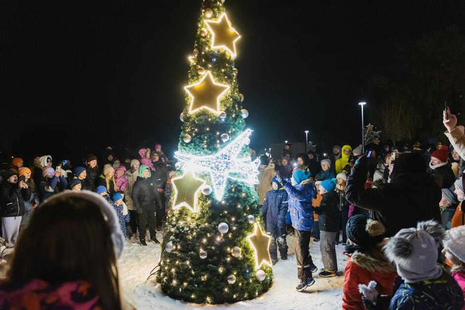 rozświetlona kilkumetrowa sztuczna choinka w nocy, na śniegu, wokół kilkadziesiąt osób, głównie małych dzieci