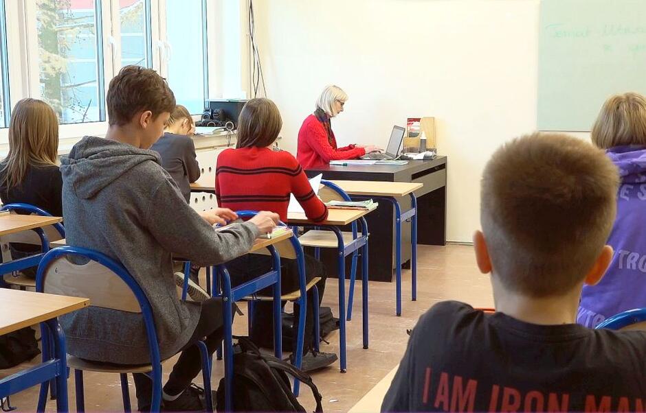uczniowie w klasie siedzą w ławkach, za nimi przy swoim stole siedzi pani nauczycielka