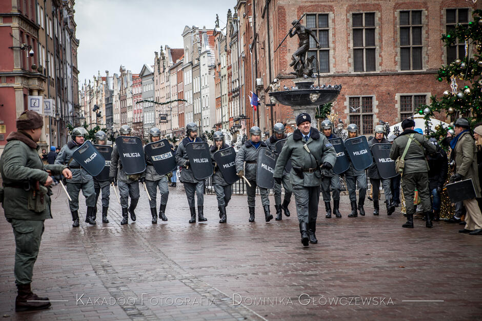 na zdjęciu grupa kilkunastu mężczyzn ubrana w dawne stroje milicyjne w raz z uzbrojeniem, idą w szeregu, widać w tle kamienice na długim targu w Gdańsku