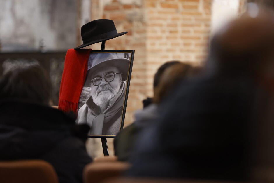 Pomieszczenie: trumna, przy niej zdjęcie mężczyzny, na który powieszone są czarny kapelusz i czerwony szal