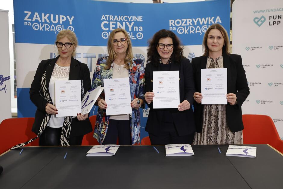 Cztery kobiety w średnim wieku stoją jedna obok drugiej, pozują do zdjęcia - każda z nich trzyma przed sobą taki sam egzemplarz umowy o współpracy, którą przed chwilą podpisały 