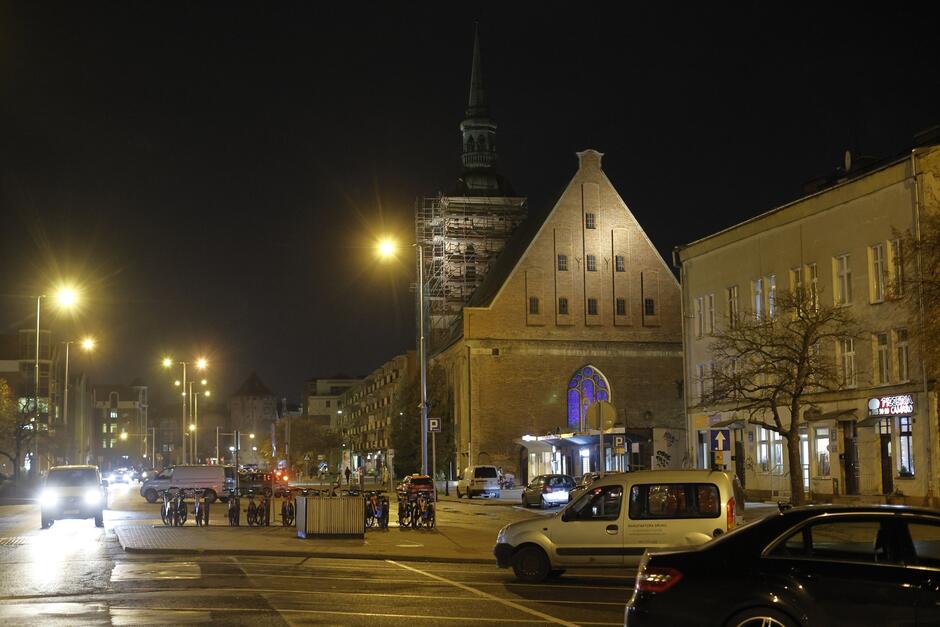 zdjęcie po zmroku, widać fragment ulicy, zaparkowane po środku rowery i kilka samochodów, po prawej widać fragmenty niewysokich budynków, w tym kościół