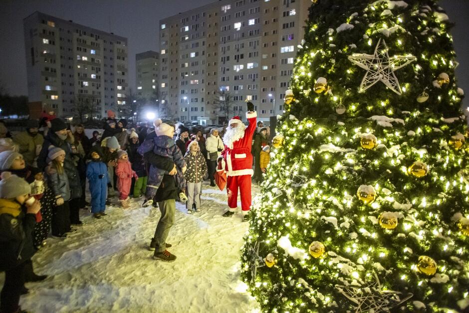 na zdjęciu z prawej rozświetlona udekorowana choinka, po lewej widać kilkanaście stojących obok siebie osób patrzących na choinkę, pomiędzy nimi Mikołaj w czerwonym stroju