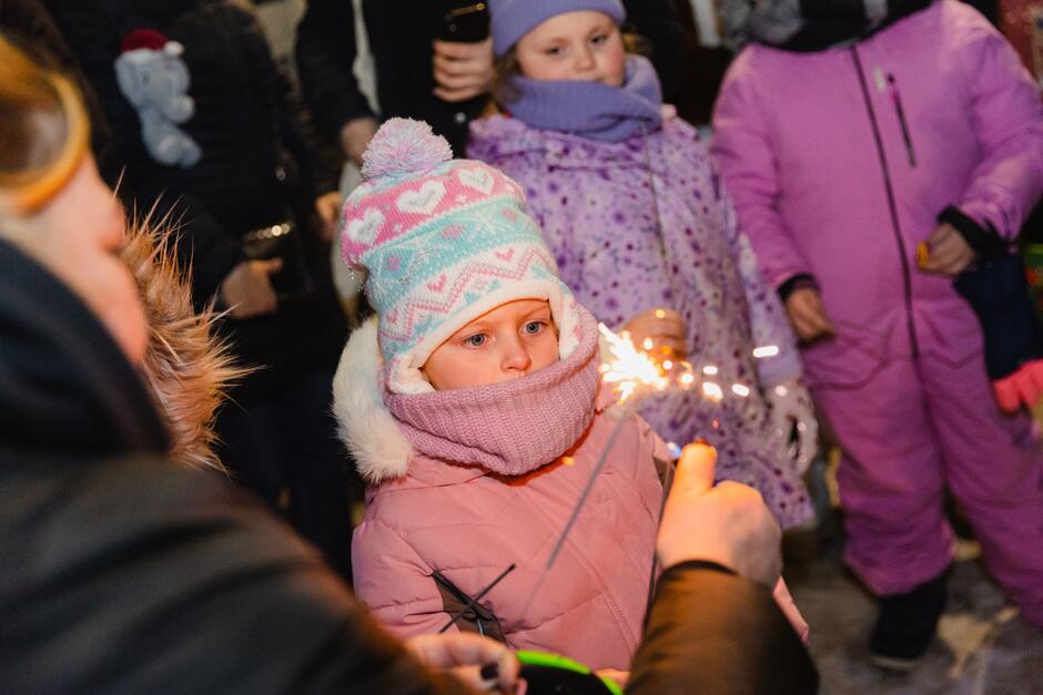 na zdjęciu kobieta w średnim wieku kuca, trzyma w ręku palce się zimne ognie, przygląda im się mała dziewczynka w ciepłym zimowym kombinezonie
