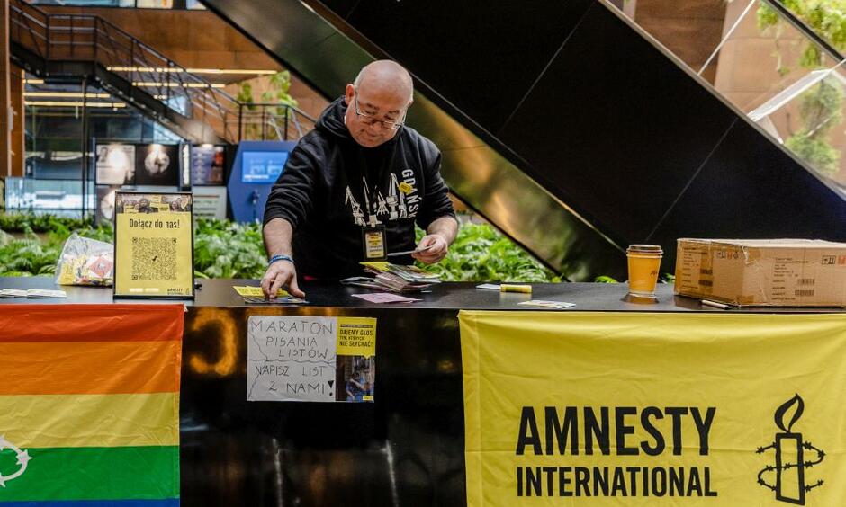 mężczyzna w średnim wieku w czarnej bluzie, pochyla się nad stolikiem na którym widać leżące ulotki, przy stoliku wisi żółty niewielki baner z napisem amnesty international, jest też zwisająca ze stolika tęczowa flaga