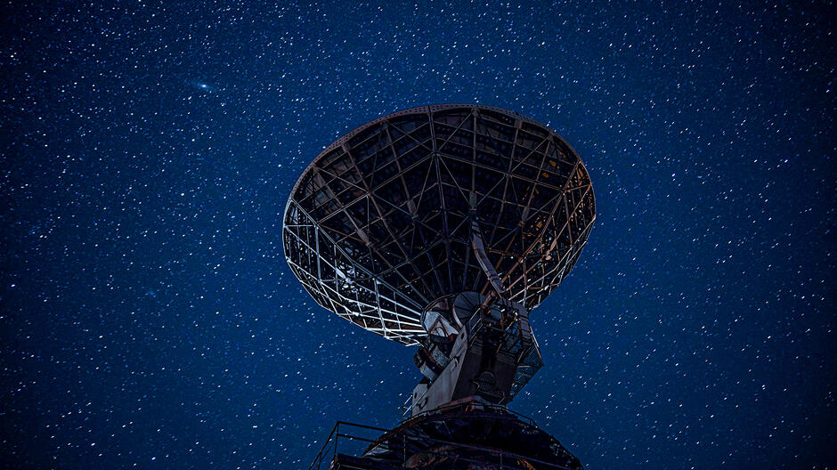 Na zdjęciu widać antenę satelitarną na tle rozgwieżdżonego nocnego nieba. Antena jest skierowana w górę, a jej powierzchnia odbija światło gwiazd, tworząc jasny punkt na zdjęciu. 
