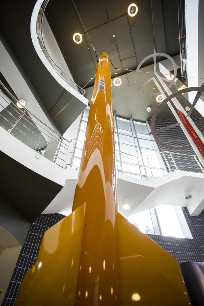 Na zdjęciu widać żółtą rakietę wiszącą z sufitu budynku. Rakieta jest skierowana w górę, a jej ogon jest podłączony do systemu dźwigów. Rakieta jest prawdopodobnie modelem, który jest używany do celów edukacyjnych lub rozrywkowych