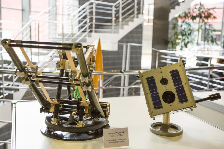 Na zdjęciu znajduje się model satelity, który spoczywa na stole. Model jest wykonany z metalu i ma kształt sześcianu. Na bokach modelu znajdują się różne oznakowania, w tym logo Europejskiej Agencji Kosmicznej (ESA) i nazwa satelity, Rosetta 