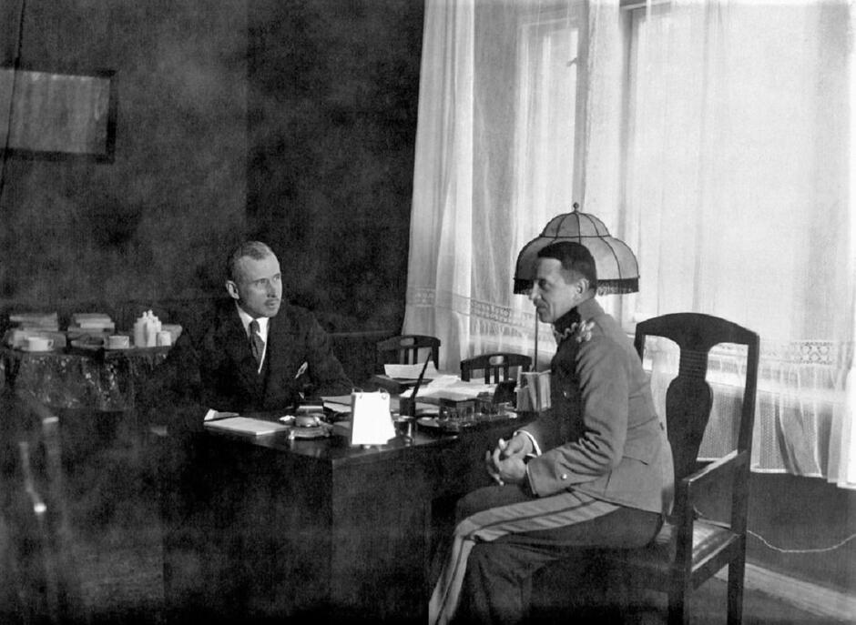 czarno białe zdjęcie przedwojenne przedstawiające dwóch mężczyzn przy biurku, jeden w mundurze, bohater opisu - w garniturze