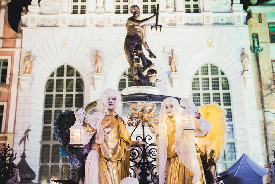 kobieta i mężczyzna przebrani za anioły na szczudłach, trzymają w dłoniach lampiony, w tle fontanna z Neptunem