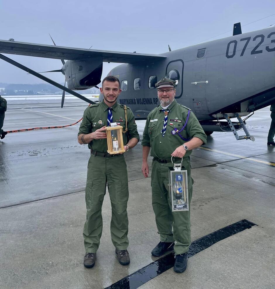 dwóch mężczyzn w mundurach harcerskich na betonowej płycie lotniska, w tle samolot wojskowy, opisany w tekście