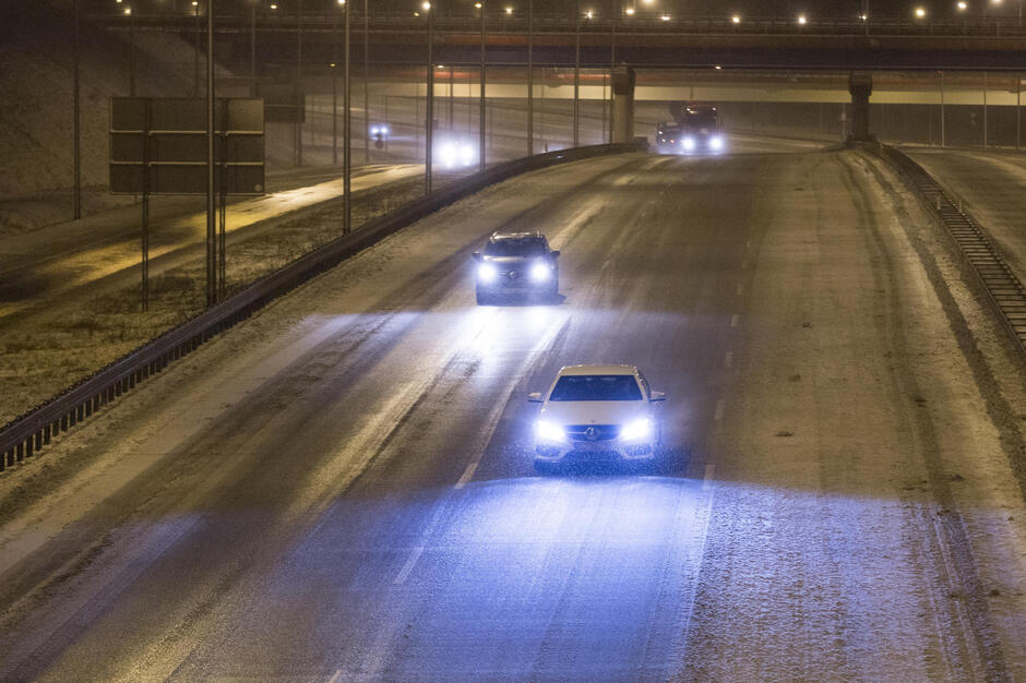 na zdjęciu ośnieżona jezdnia, zdjęcie wykonane wieczorem widać świecące latarnie przy ulicy i kilka jadących samochodów