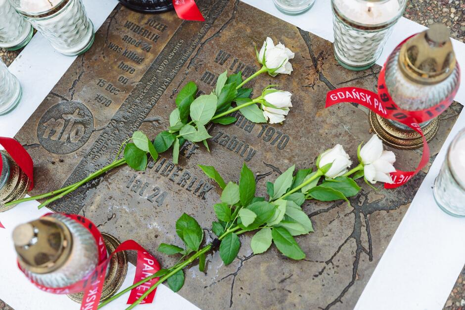 Tablica z brązu z napisami, które są częściowo zasłonięte przez znicze, kwiaty i biało-czerwone wstążki