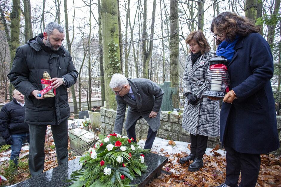 Cztery osoby z kwiatami i zniczami, stoją przy grobie na cmentarzu