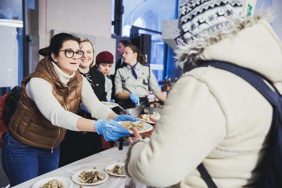 na zdjęciu prezydent gdańska, kobieta w średnim wieku w jasnobrązowej kamizelce i rękawiczkach jednorazowych w kolorze niebieskim podaje plastikowy talerz z jedzeniem kobiecie ubranej w zimowe jasne ciuchy