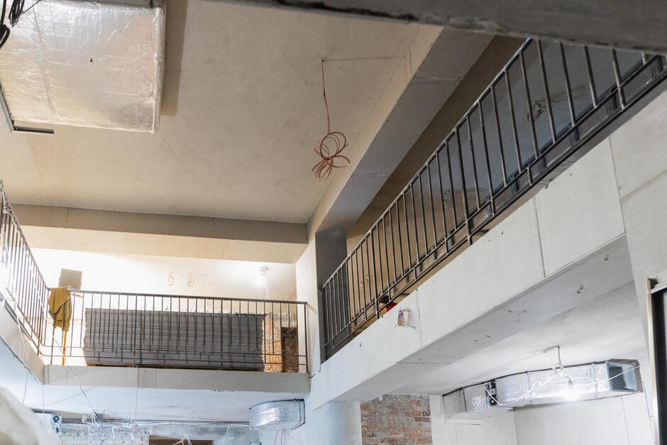 Na zdjęciu widać balkon na drugim piętrze budynku w budowie. Balkon jest wykonany z betonu i metalu. Ma balustradę z metalowych prętów. Balkon jest pusty, nie ma na nim żadnych mebli ani roślin