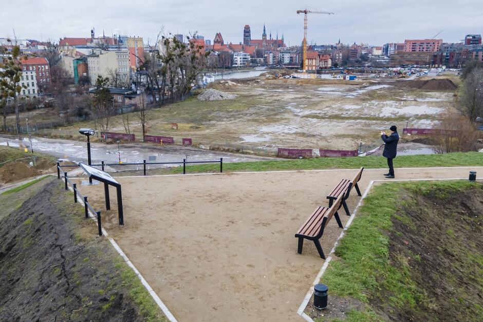 zdjęcie z drona, widać placyk z nawierzchni piaskowej przy nim kilka ławek po prawej, po lewej widać lunetę, w tle plac budowy