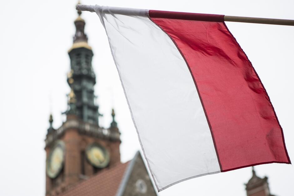 na zdjęciu na pierwszym planie biało-czerwona flaga, w tle fragment zabytkowego budynku kościoła z tarczą zegarową