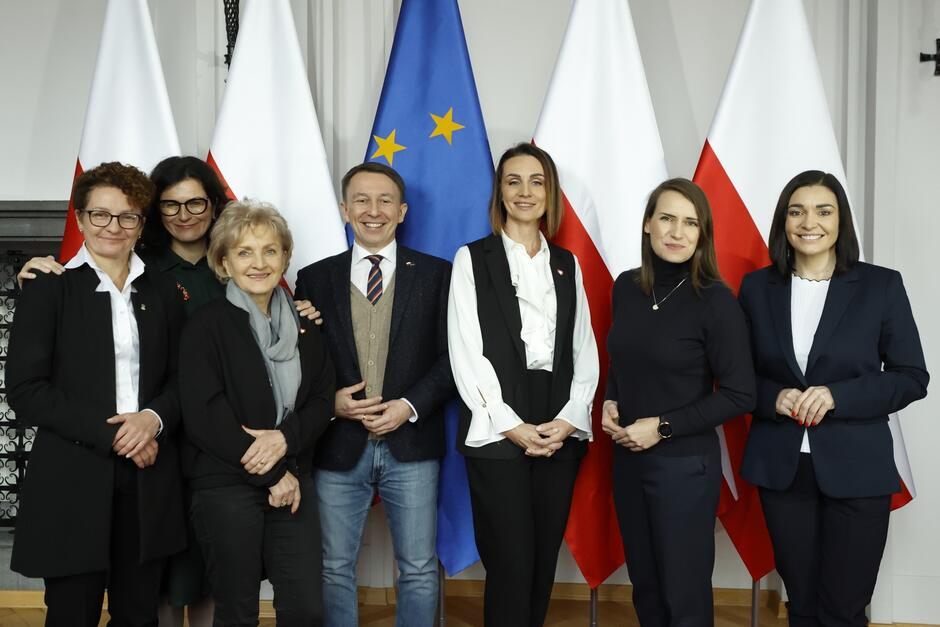 Kilka kobiet i mężczyzna pozujący do wspólnego zdjęcia na tle flag Polski i Unii Europejskiej