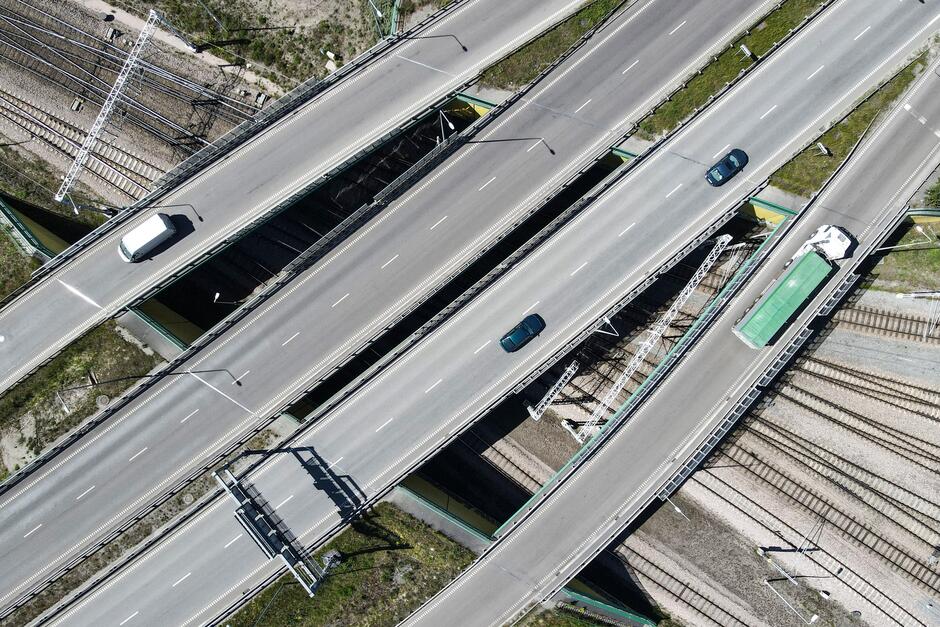 Zdjęcie przedstawia z lotu ptaka skrzyżowanie dwóch poziomów autostrady. Widoczne są dwa poziomy wiaduktów: jeden przebiega poziomo przez całe zdjęcie, a drugi przechodzi pod nim tworząc krzyż. Na obu poziomach drogi znajdują się samochody: na górnym poziomie są trzy, a na dolnym jeden zielony autobus. Poniżej i po bokach autostrad widać tory kolejowe z kilkoma masztami sieci trakcyjnej, co wskazuje na bliskość infrastruktury kolejowej