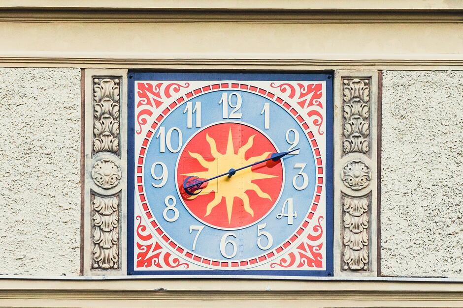 na zdjęciu zbliżenie na zegar umieszczony na budynku, widać cyfry arabskie, jedna czarną wskazówkę, czerwone i niebieskie tło