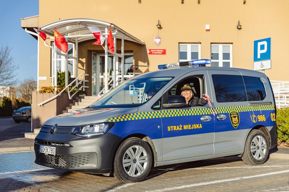 na zdjęciu radiowóz straży miejskiej, oznakowany, za kierownicą siedzi strażniczka z blond włosami, wystawia rękę przez otwarte okno pokazując klucze, w tle niewysoki budynek komendy