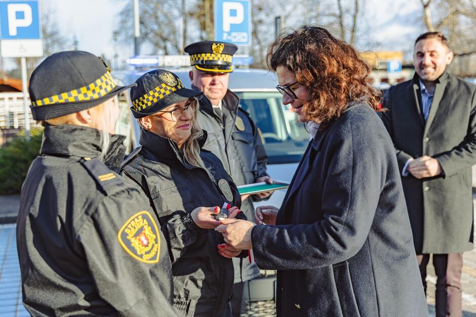 na zdjęciu prezydent gdańska w granatowym płaszczu po prawej rozmawia z dwiema strażniczkami miejskimi które stoją po lewej, w tle dwóch mężczyzn
