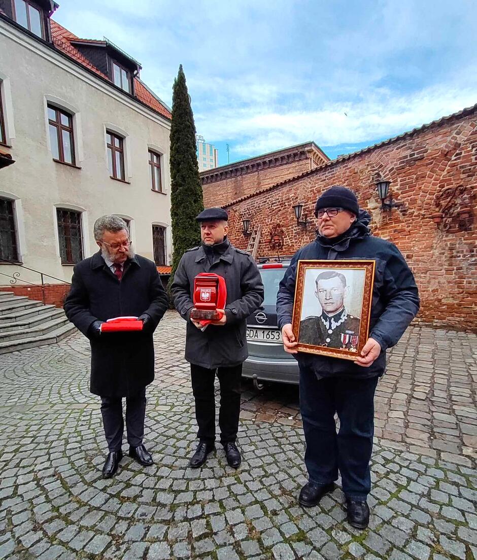 Zdjęcie przedstawia trzech mężczyzn stojących na brukowanym dziedzińcu przed budynkiem o tradycyjnej architekturze. Każdy z mężczyzn trzyma przedmiot związany z ceremonią lub wydarzeniem pamiątkowym. Mężczyzna po lewej stronie trzyma czerwoną urnę z godłem Polski, mężczyzna po prawej stronie trzyma oprawiony w ramkę portret mężczyzny w mundurze, a osoba w środku trzyma niewidoczny dla nas przedmiot. Wszyscy są ubrani w zimowe okrycia wierzchnie, co wskazuje na chłodną porę roku
