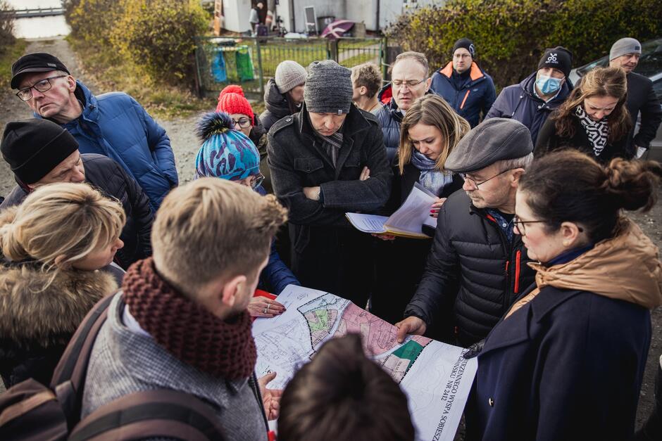 na zdjęciu grupa kilkunastu osób, skupionych w okręgu, w środku osoba trzymająca papierową mapę, inni się jej przyglądają