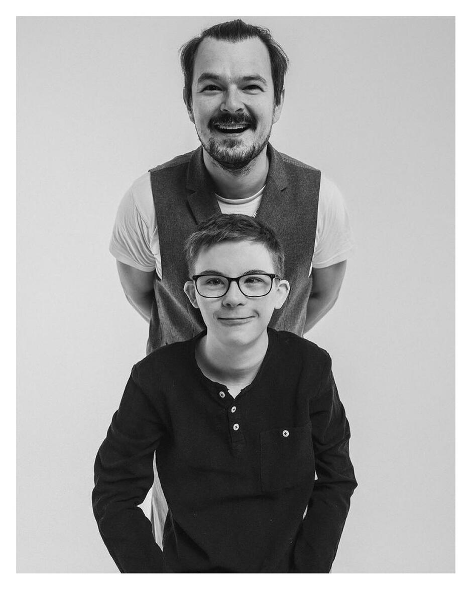 Czarno-białe zdjęcie: mężczyzna z zarostem, przed nim (głowę niżej) chłopiec z zespołem Downa. Uśmiechają się 