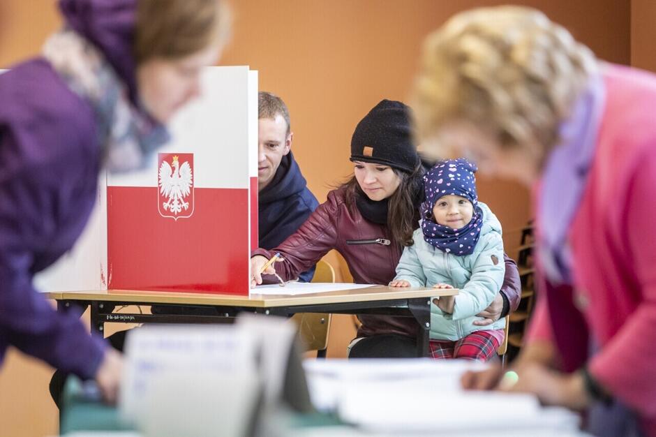 Na pierwszym planie (zamazana) kobieta stoi nad kartką, w tle inna kobieta z dzieckiem patrzącym w obiektyw siedzi przy stoliku