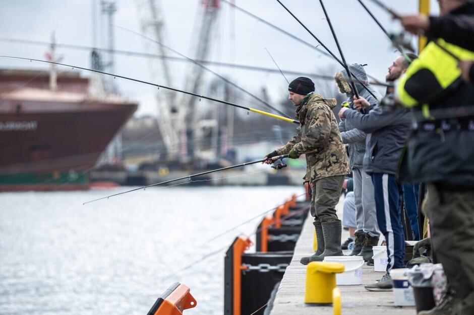 Wędkarze stoją z zarzuconymi wędkami nad rzeką. W tle statek i dźwigi portowe