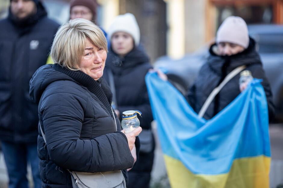 Płacząca kobieta w kurtce trzyma palący się znicz, w tle ille osoby, w tym z flagą Ukrainy