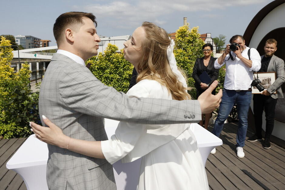 młoda para w ślubnych strojach całuje się, za nimi fotograf robi zdjęcia, stoi kobieta w średnim wieku i młody mężczyzna