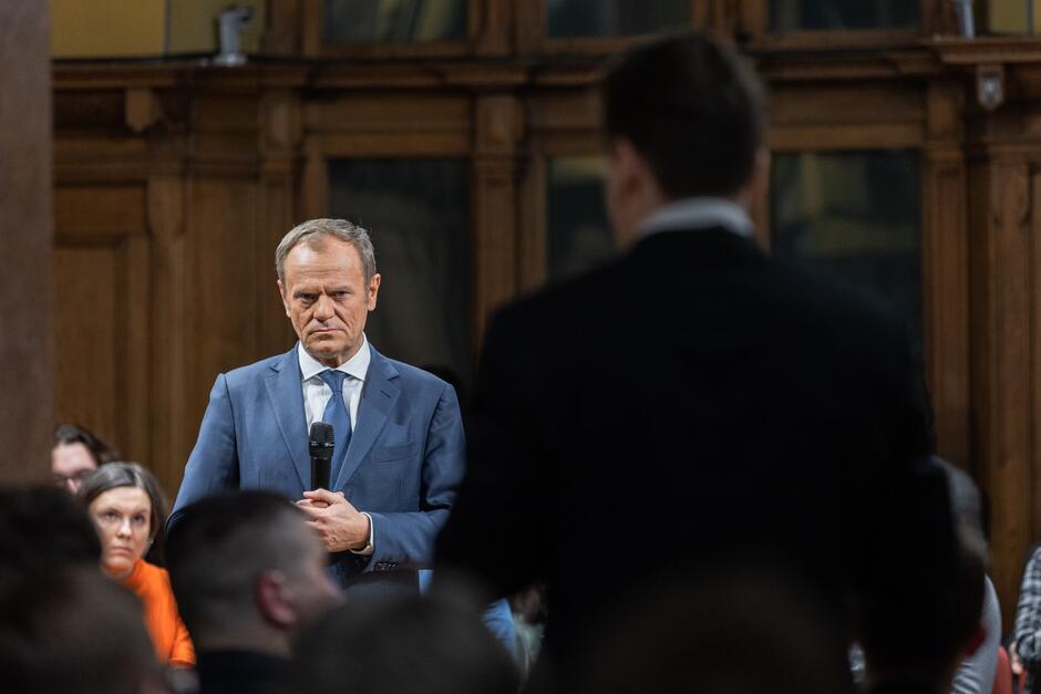 Zdjęcie przedstawia Donalda Tuska na środku sali, otoczonego uczestnikami spotkania