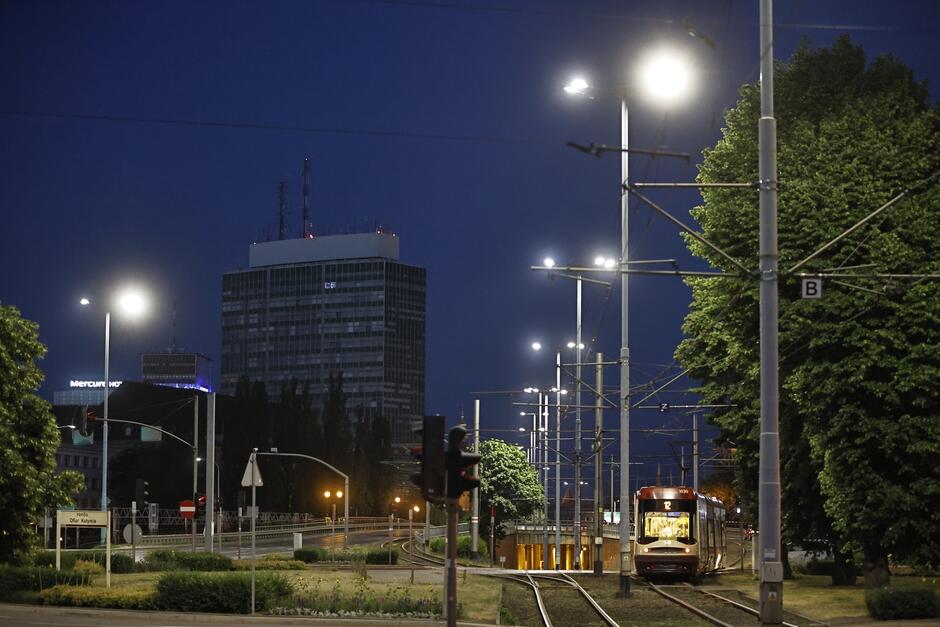 zdjęcie wykonane po zmroku, widać fragment ulicy ze świecącymi się przy niej latarniami, w tle wysoki budynek biurowca na kilkadziesiąt pięter