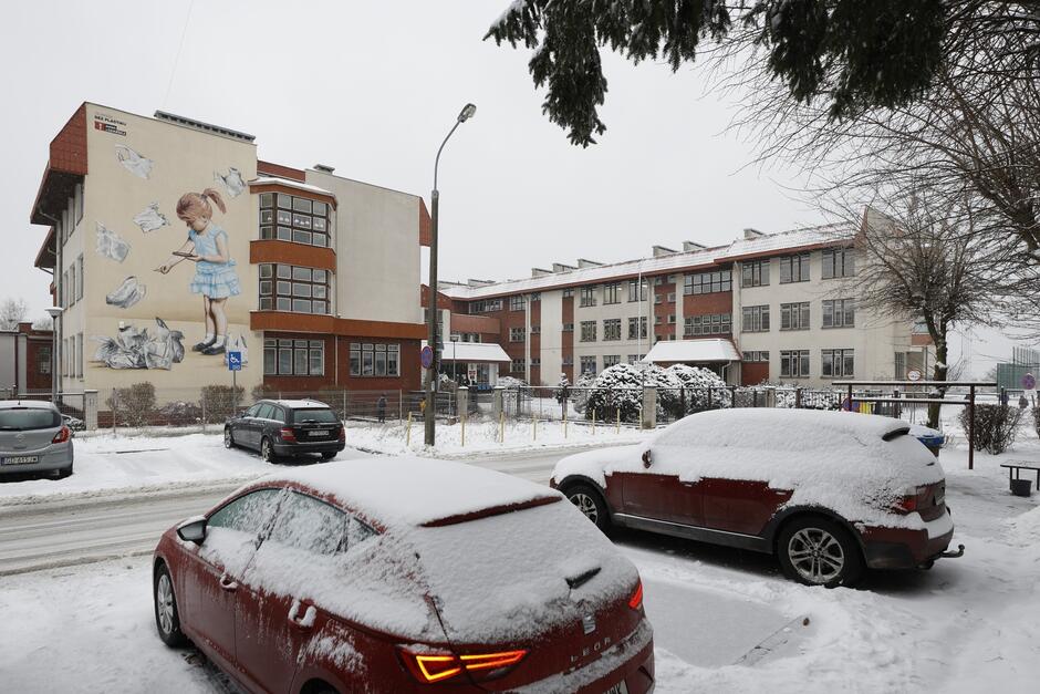 na pierwszym planie dwa pokryte śniegiem samochody osobowe, zaparkowane przed dwuskrzydłowym kilkupiętrowym budynkiem szkolnym, też pokrytym śniegiem