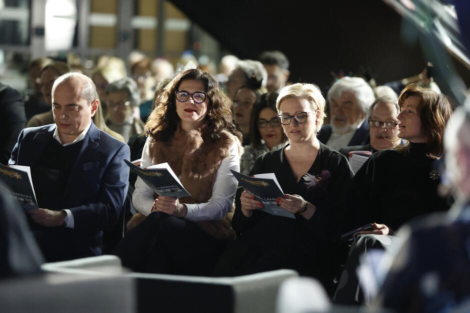 Sala z widownią. Na pierwszym planie (od lewej) mężczyzna, kobieta w okularach i kobieta, trzymają w dłoniach śpiewniki 