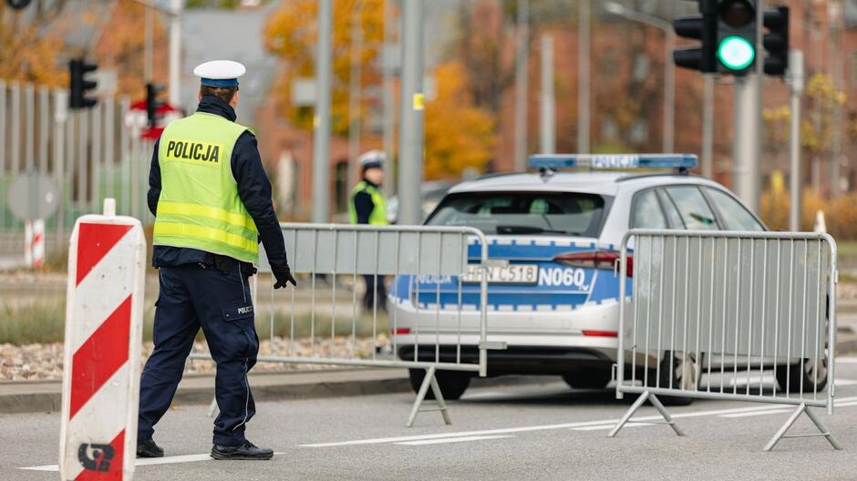 Na zdjęciu widzimy scenę z miejskiego otoczenia, gdzie policjant w niebieskim mundurze i jaskrawo-żółtej kamizelce odblaskowej kieruje ruchem. Obok niego jest biała barierka drogowa z czerwonymi paskami. W tle widoczny jest radiowóz policyjny z charakterystycznymi niebiesko-białymi barwami oraz napisem POLICJA  na boku. Scena wygląda na kontrolę lub blokadę drogową. Na ulicy widoczne są sygnalizatory świetlne, z których jeden świeci na zielono. Za barierkami dostrzec można drugiego policjanta