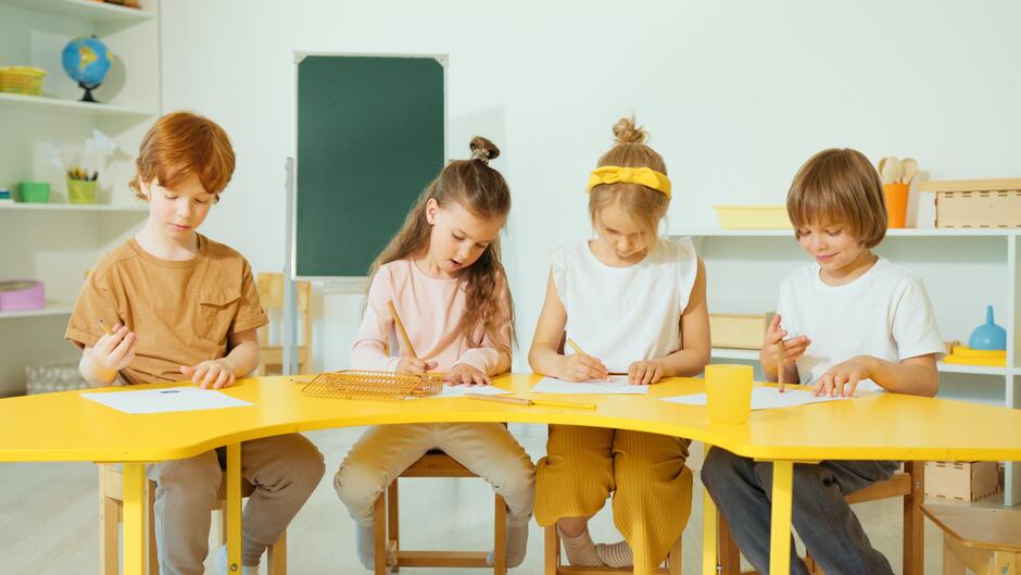 Na zdjęciu widzimy czworo dzieci siedzących przy żółtym stole i skupionych na pracy z papierem i kredkami. Każde dziecko jest zanurzone w swojej aktywności twórczej, rysując lub pisząc. W tle widzimy klasę szkolną z tablicą, globusem i regałami z różnymi materiałami edukacyjnymi. 