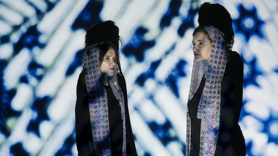 Na zdjęciu widać dwie kobiety w ozdobnych strojach, stojące na tle projekcji z abstrakcyjnym, geometrycznym wzorem. Mają czarne ubrania, a na głowach mają długie szale ze srebrnymi elementami. 