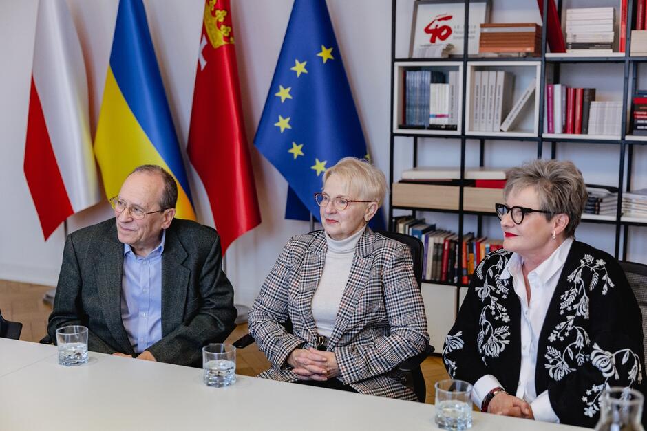 na zdjęciu dwie starsze kobiety i jeden starszy mężczyzna, elegancko ubrani, siedzą za stołem, w tle za nimi regał z książkami i flagi gdańska, polski oraz unii europejskiej