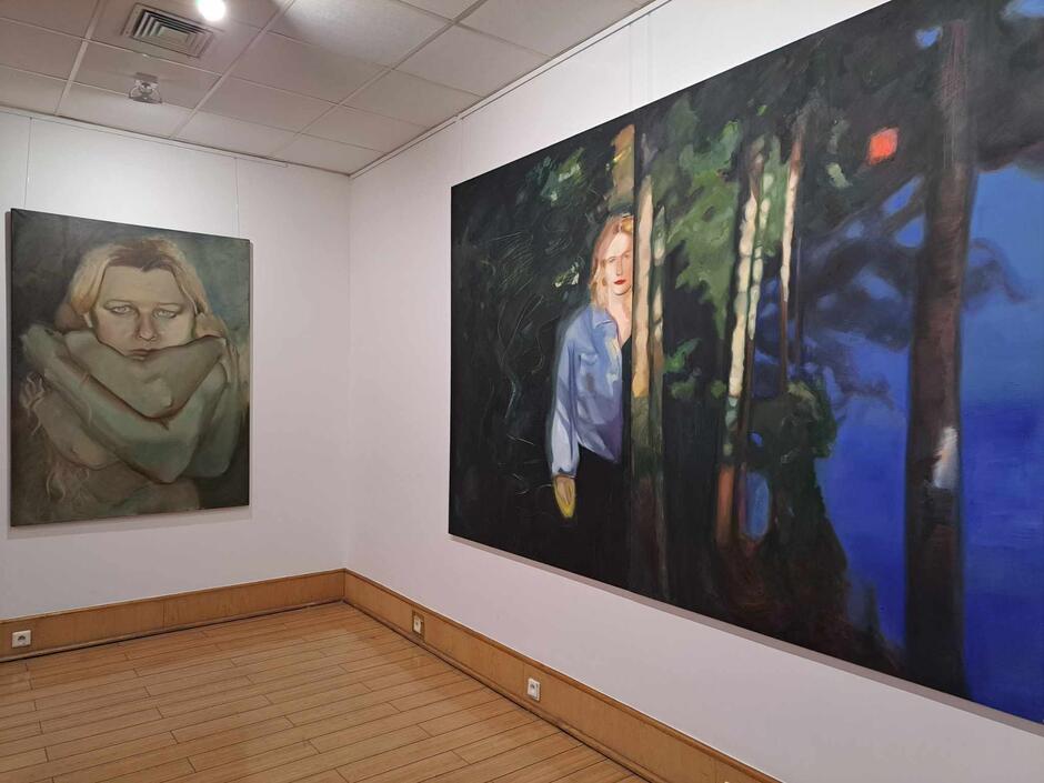 Na zdjęciu widzimy wnętrze galerii z dwoma dużymi obrazami. Po lewej stronie znajduje się portret kobiety oplecionej ramionami, z zamyślonym wyrazem twarzy, a po prawej większe płótno przedstawiające stojącą kobietę na tle lasu i jeziora w nocy.