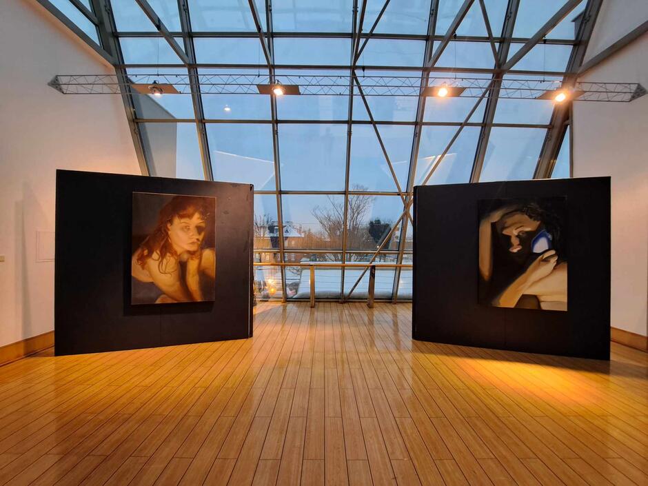 Na zdjęciu znajduje się wnętrze galerii sztuki z dużymi oknami, przez które widać zmierzch. Prezentowane są dwa duże portrety na czarnych ścianach wystawienniczych, oba przedstawiające kobiety; jeden z obrazów pokazuje kobietę trzymającą telefon przy uchu.