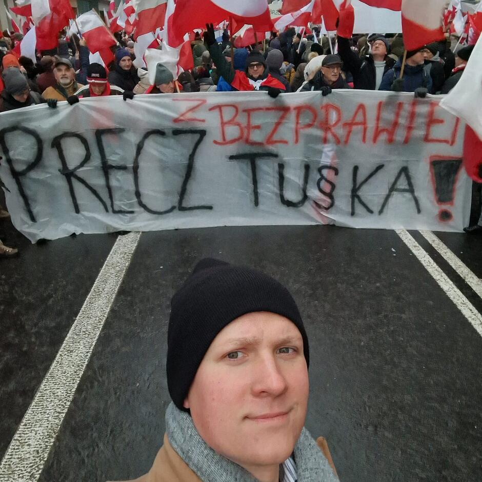 Młody mężczyzna we włóczkowej czapce robi sobie selsie - w tle tłum ludzi z flagami narodowymi i transparent: Precz z bezprawiem Tuska 