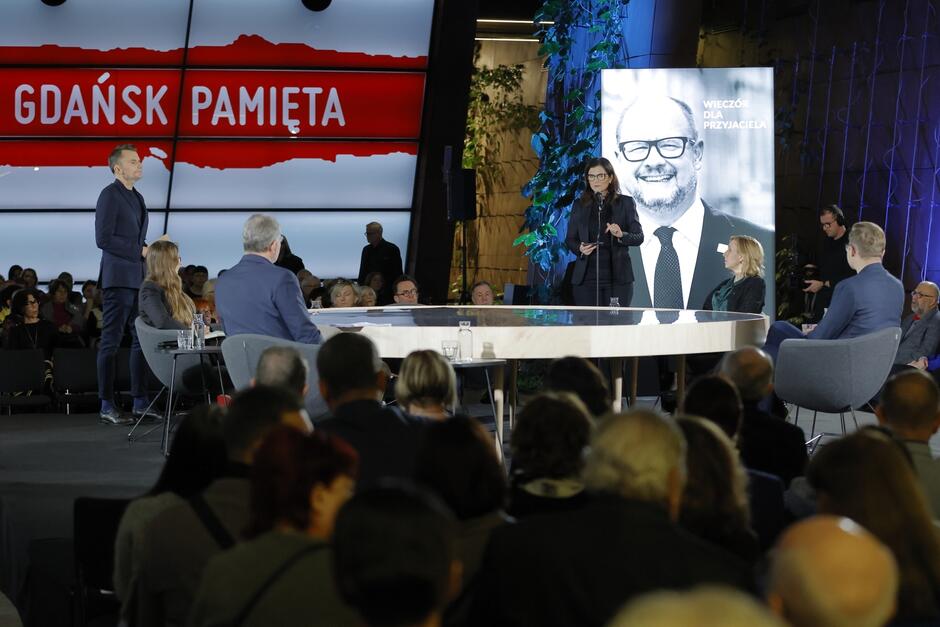 na zdjęciu na podwyższeniu stoi okrągły duży stół przy którym siedzi pięć osób, obok stoi kobieta w średnim wieku w granatowej sukience, widać też duże czarno białe zdjęcie prezydenta pawła adamowicza