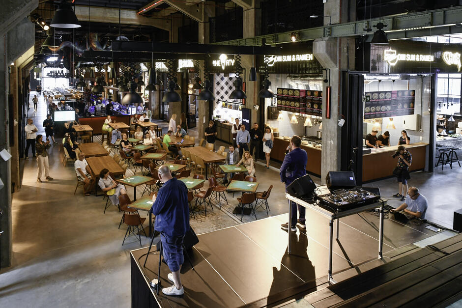 industrialna przestrzeń, w niej stoliki i krzesła, w tle restauracje i bary, na pierwszym planie niewielka scena