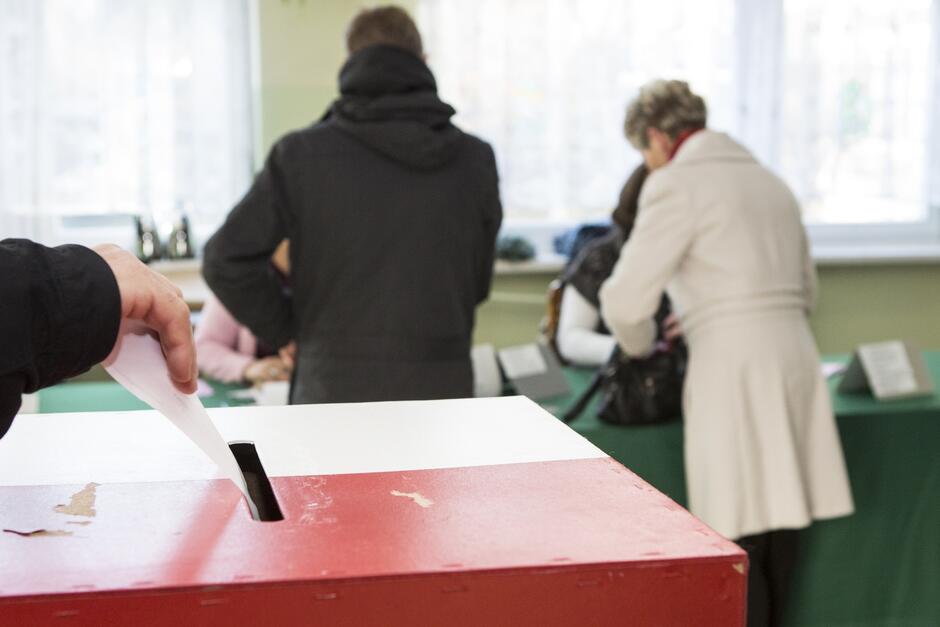 Ręka wrzucająca kartę wyborcą do urny wyborczej. W tle dwie osoby pobierają od komisji wyborczej karty do głosowania