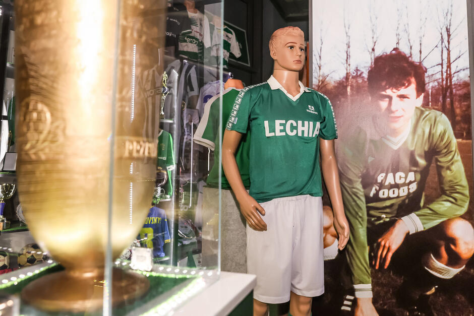 Zielona koszulka Lechii na manekinie, obok zdjęcie z kucającym mężczyzną w stroju sportowym. Na pierwszym planie po lewej puchar