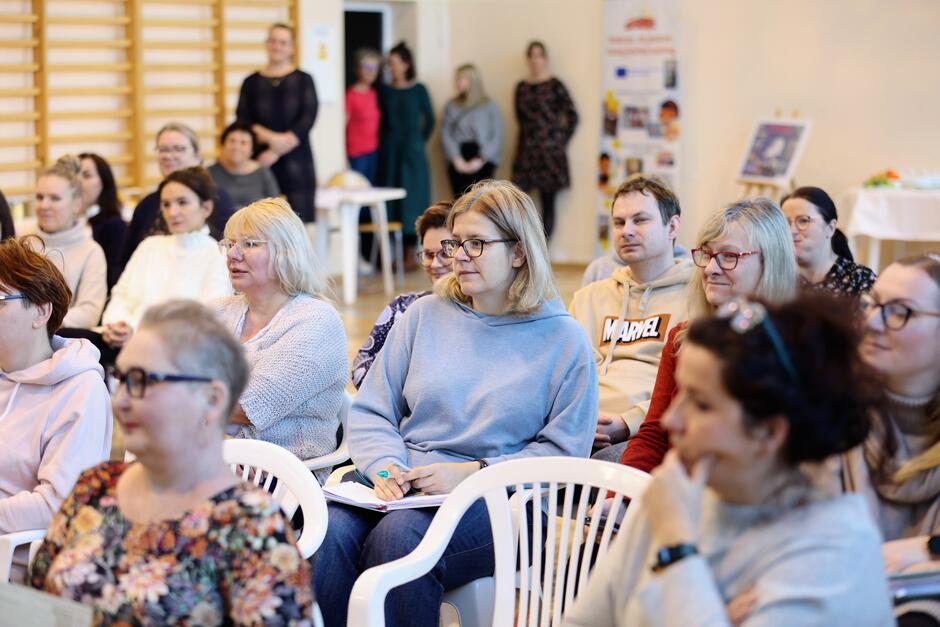 Na zdjęciu widzimy grupę ludzi siedzących na białych plastikowych krzesłach w jasnym pomieszczeniu. Wśród publiczności można dostrzec różnorodność wieku i stylów ubioru, od casualowych do bardziej formalnych. Większość osób skupia uwagę na czymś, czego nie widać na zdjęciu, prawdopodobnie na osobie mówiącej lub wydarzeniu poza kadrem. W tle, po prawej stronie, stoi roll-up z grafikami i tekstem, który wydaje się zawierać informacje związane z wydarzeniem, a na lewo od publiczności znajduje się stół z pewnymi przedmiotami, co może sugerować część cateringową lub wystawę. Na pierwszym planie widoczne są głównie kobiety, z wyjątkiem jednego mężczyzny w swetrze z napisem Marvel . Wyraz twarzy uczestników jest skupiony i zainteresowany. Atmosfera zdjęcia jest spokojna, a otoczenie wygląda na typowe dla szkolnych lub społecznych zgromadzeń. Przestrzeń z drewnianą podłogą i żółtymi ścianami sprawia wrażenie przytulności